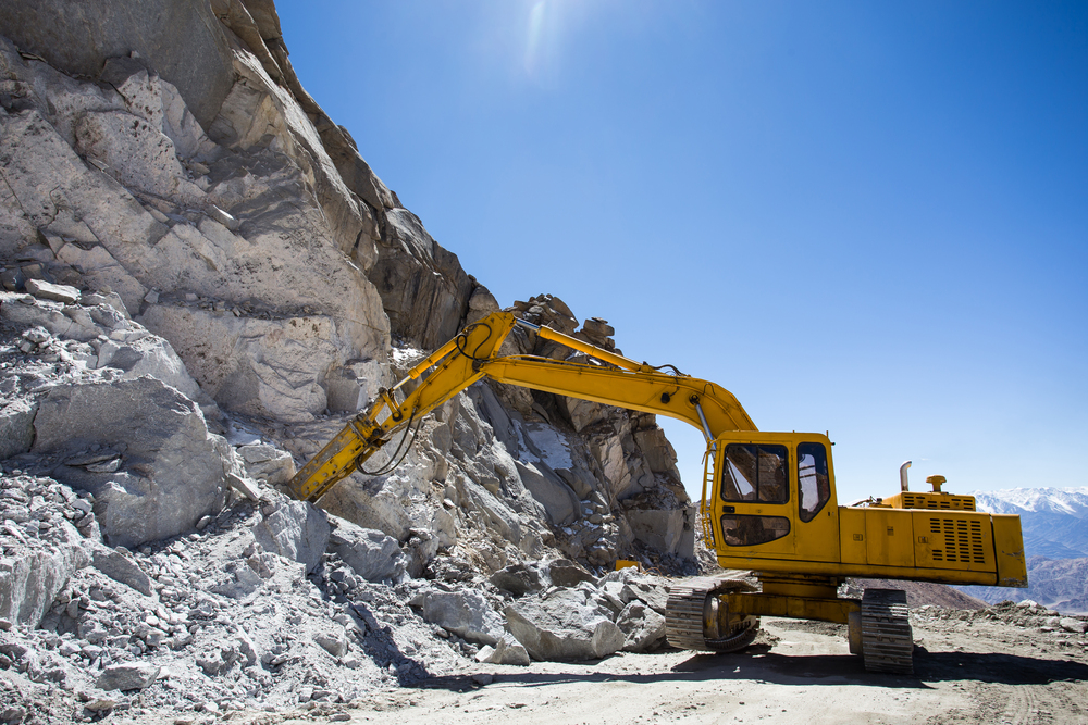 Yellow excavator performing rock excavation on a steep cliffside in Banning, CA, under clear blue skies.